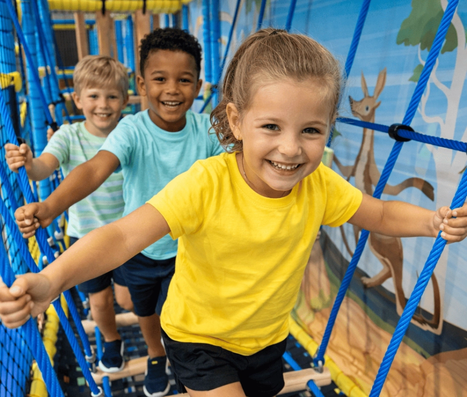 Trois enfants souriants en train de franchir un pont dans un parc de jeux indoor